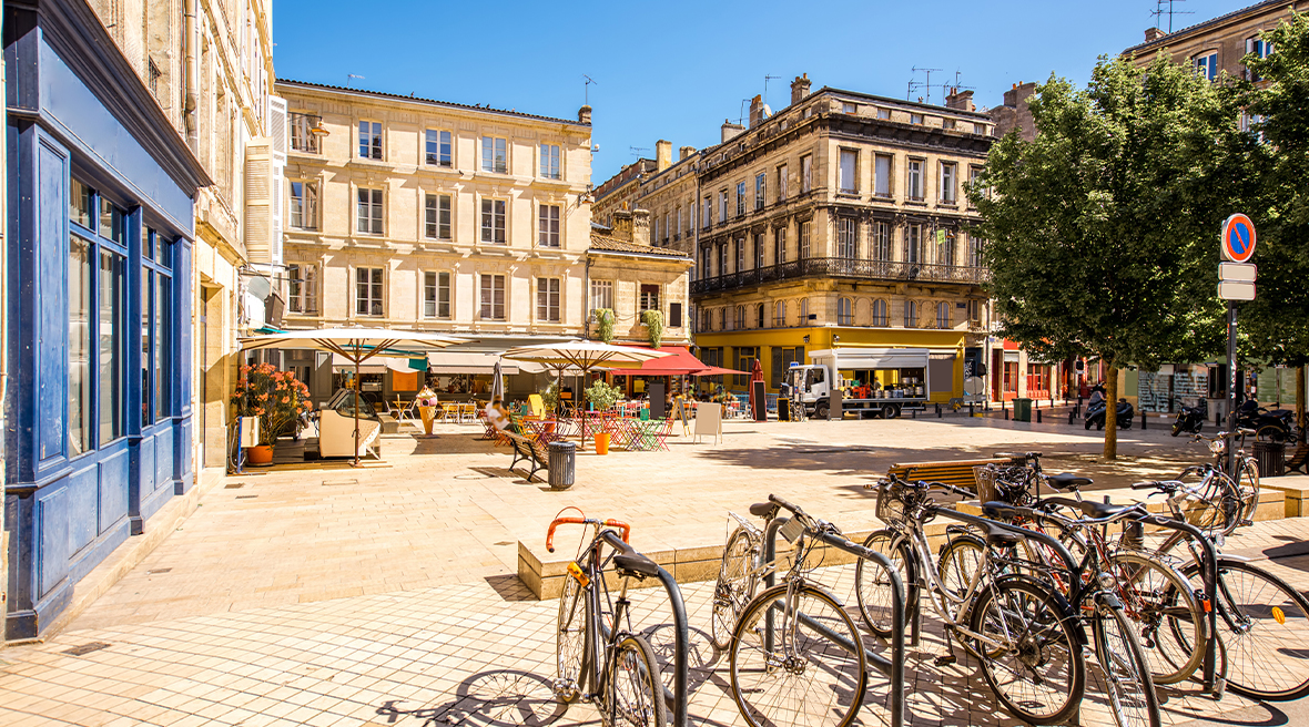 Bicycles locked up in a sunshine filled square surrounded by ornate buildings with a covered café in the square